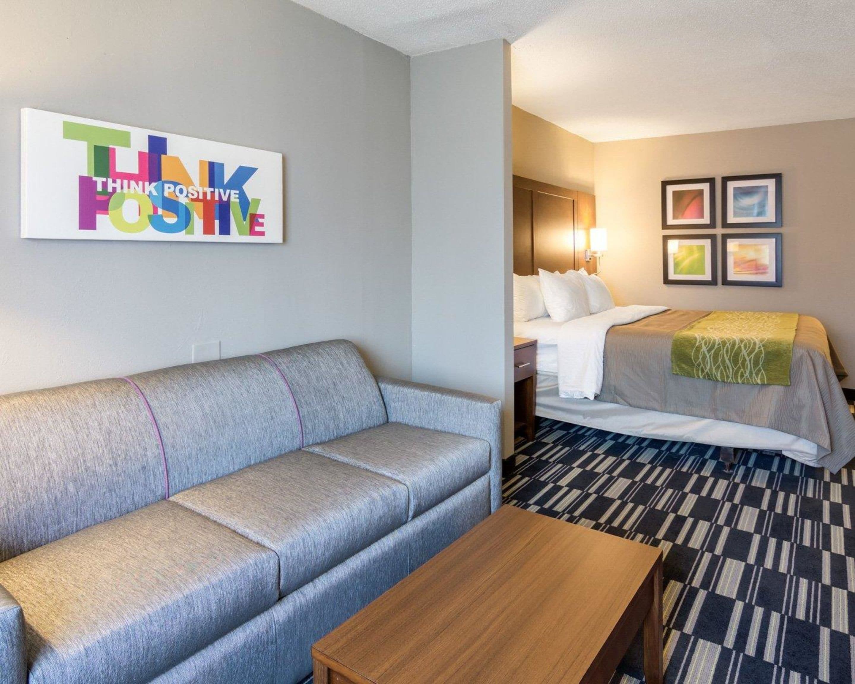 A hotel room with a grey sofa, a wooden coffee table, a bed with white bedding, and a patterned carpet. There is a wall with a 'THINK POSITIVE' sign.