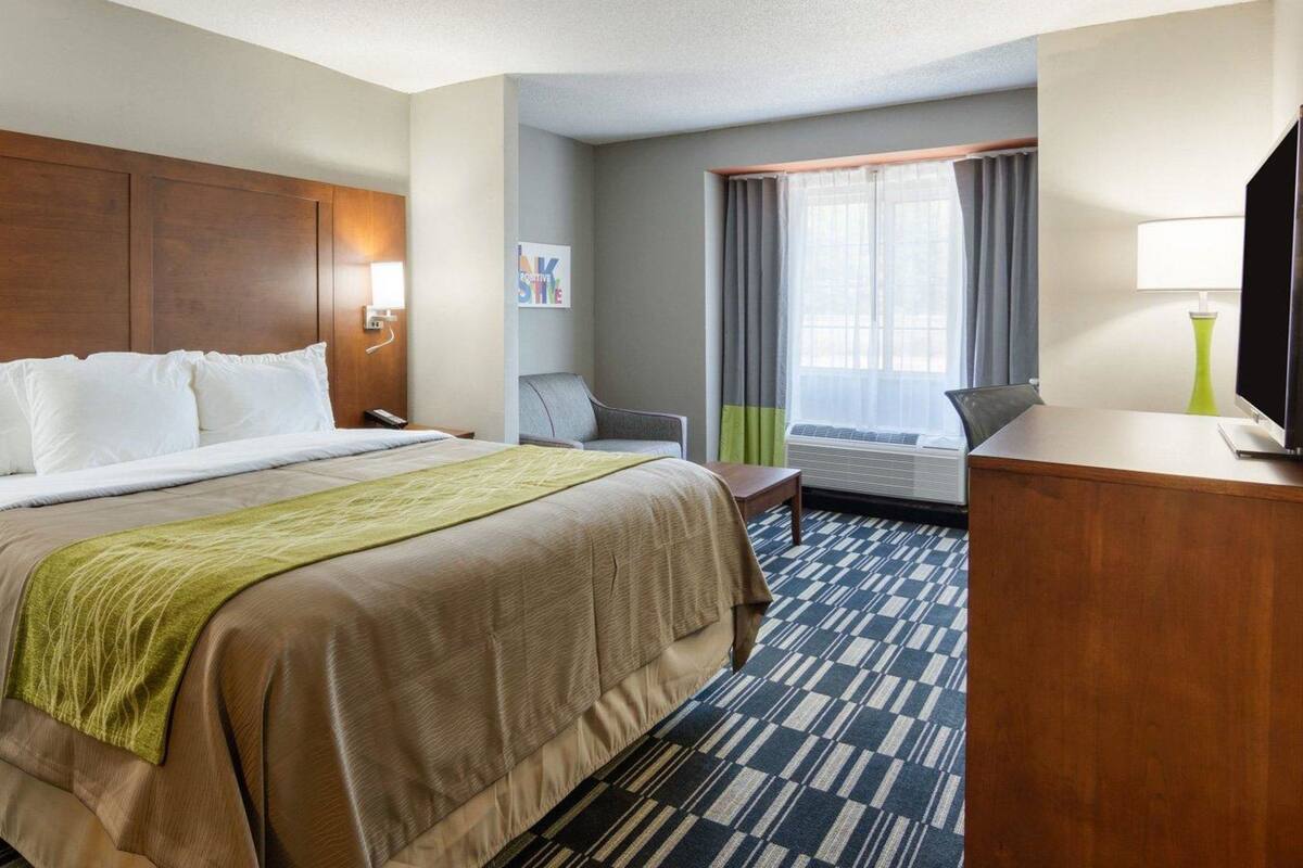 A hotel room with a large bed, a desk with a lamp, a chair, a window with curtains, and a television.
