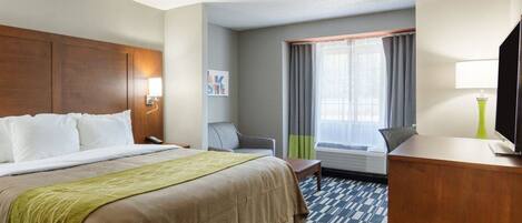 A hotel room with a large bed, a desk with a lamp, a chair, a window with curtains, and a television.