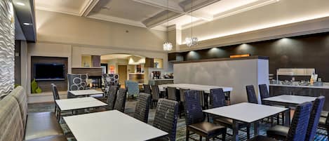A hotel dining area with multiple tables and chairs, a kitchen area, and a seating area with a sofa and a TV.