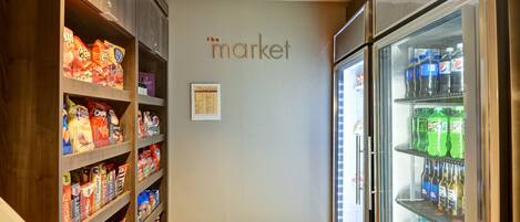 A narrow hallway with shelves stocked with snacks and beverages, a refrigerator with drinks, and a wall sign reading "the market".