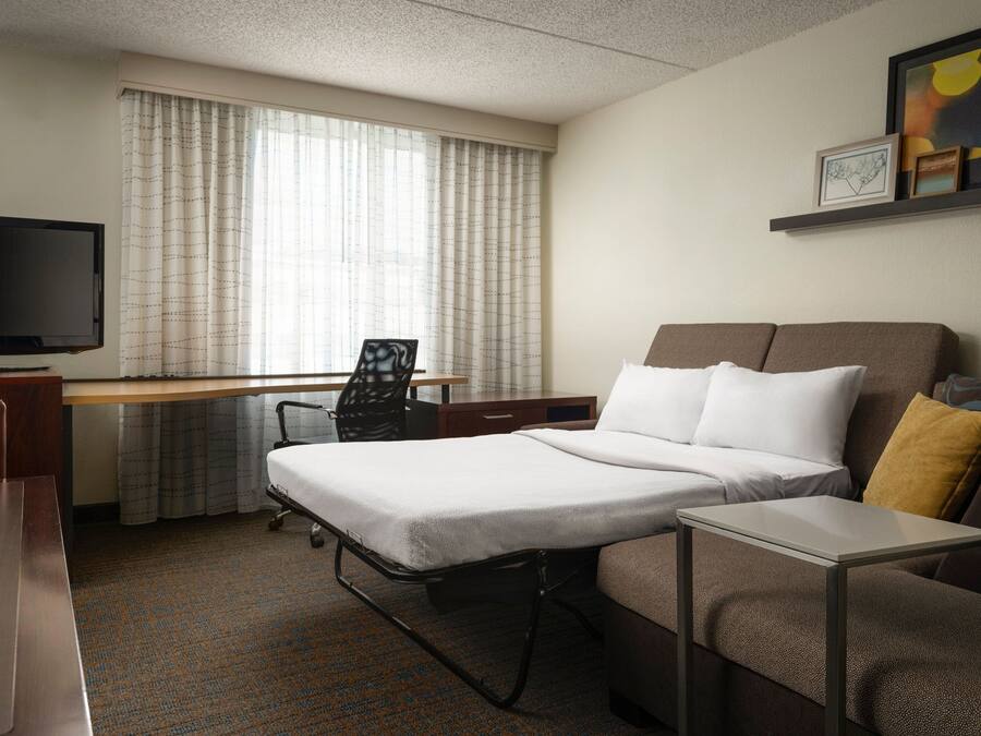 Quarto de hotel com cama, escrivaninha, cadeira, televisão e sofá.