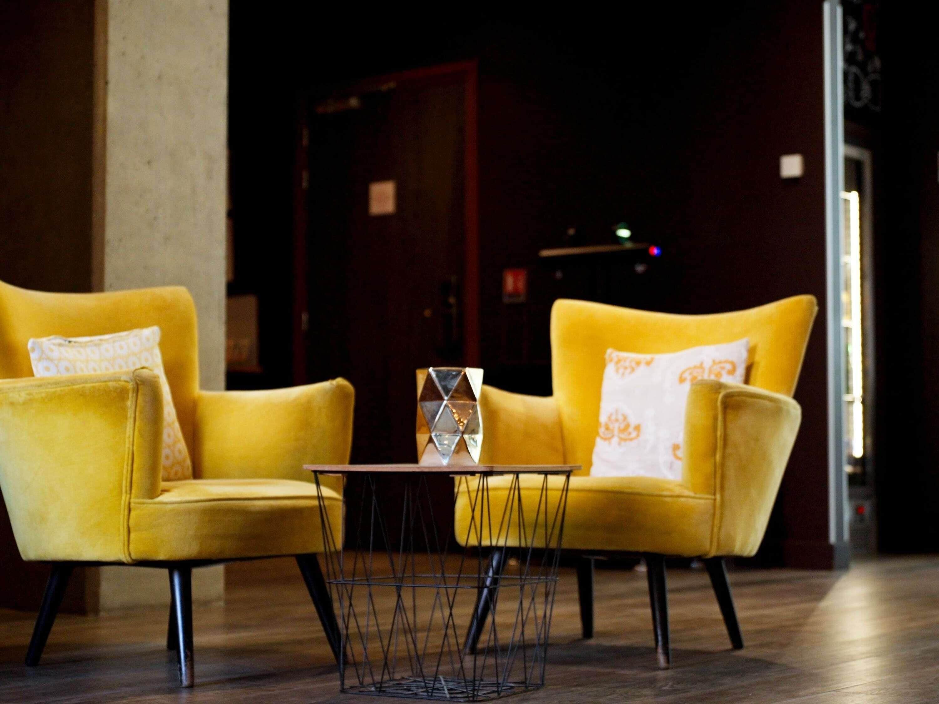 Deux fauteuils jaunes ornés de coussins à motifs, une petite table ronde surmontée d’un centre de table géométrique en verre, et un plancher en bois foncé.