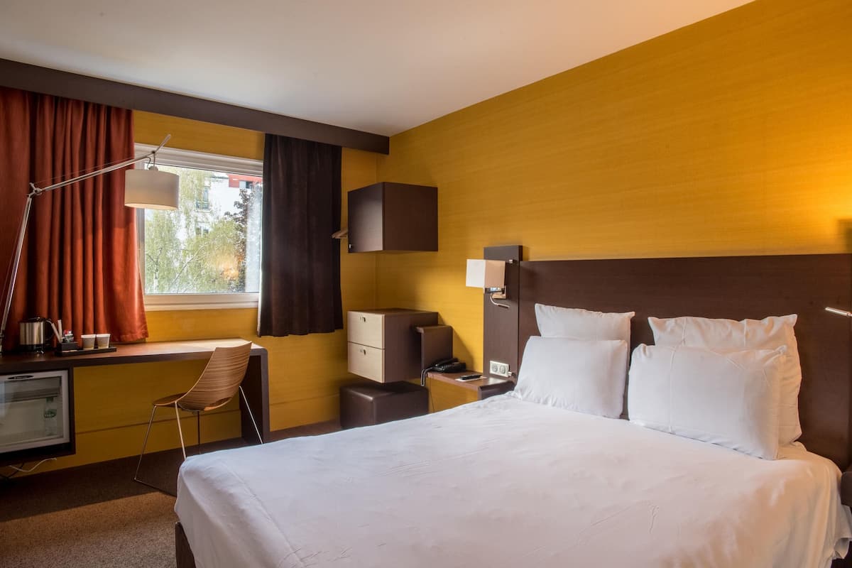 A hotel room with a large bed, a desk with a chair, a lamp, a wardrobe, and a window with curtains.