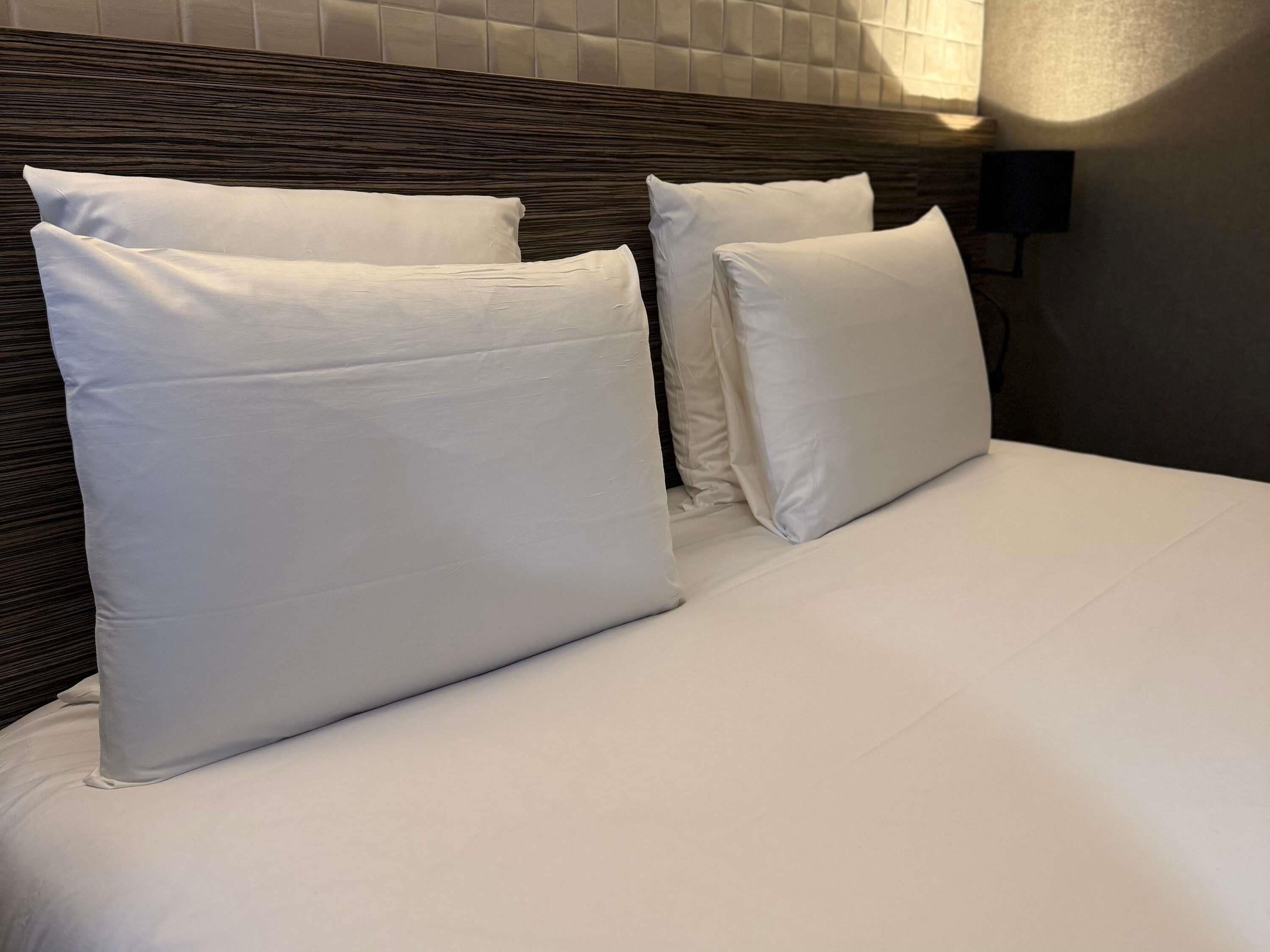 Una cama bien arreglada con sábanas y almohadas blancas, cabecera de madera oscura y aplique de pared.
