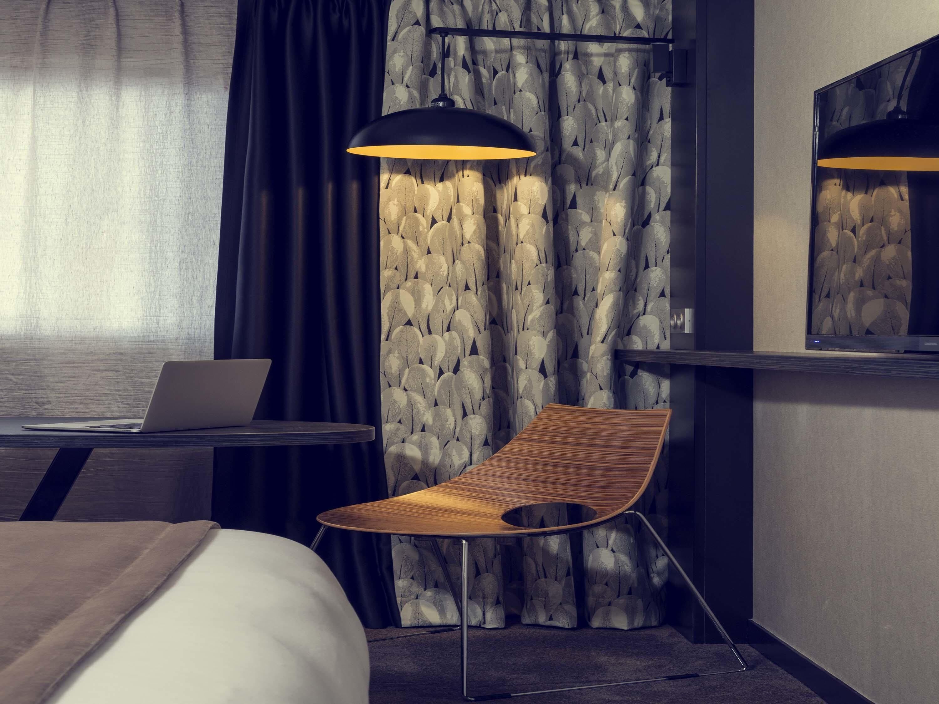 A modern hotel room with a wooden chair, a desk with a laptop, and a large mirror.
