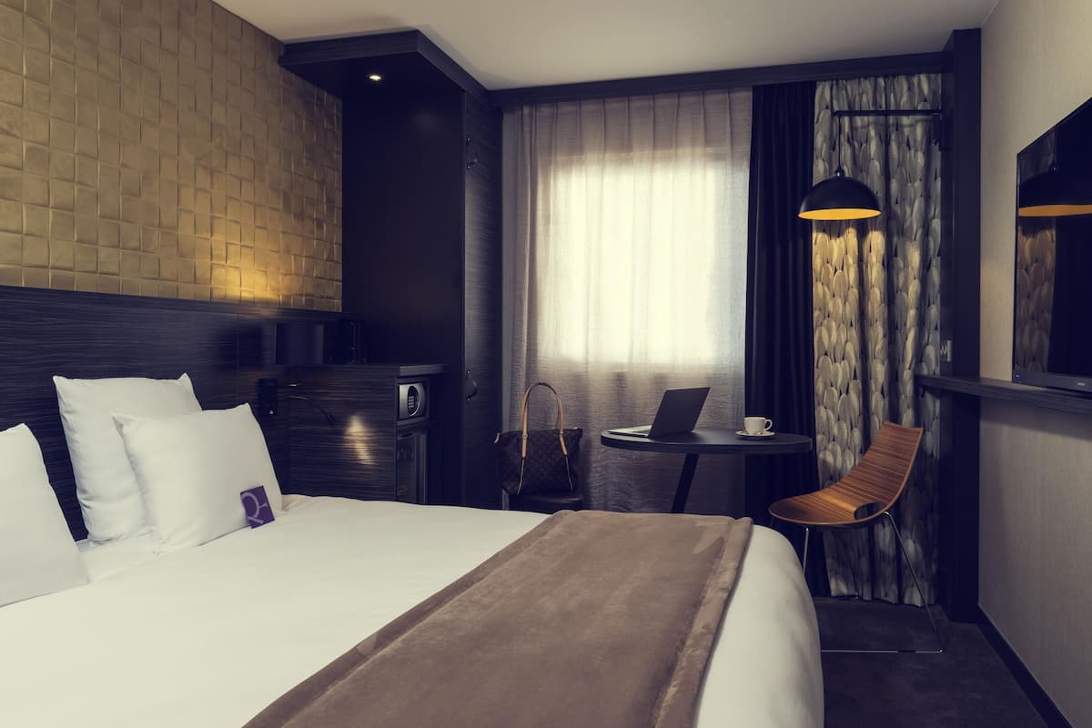 A hotel room with a large bed, a desk with a laptop, a chair, a small table with a cup, and a mirror.
