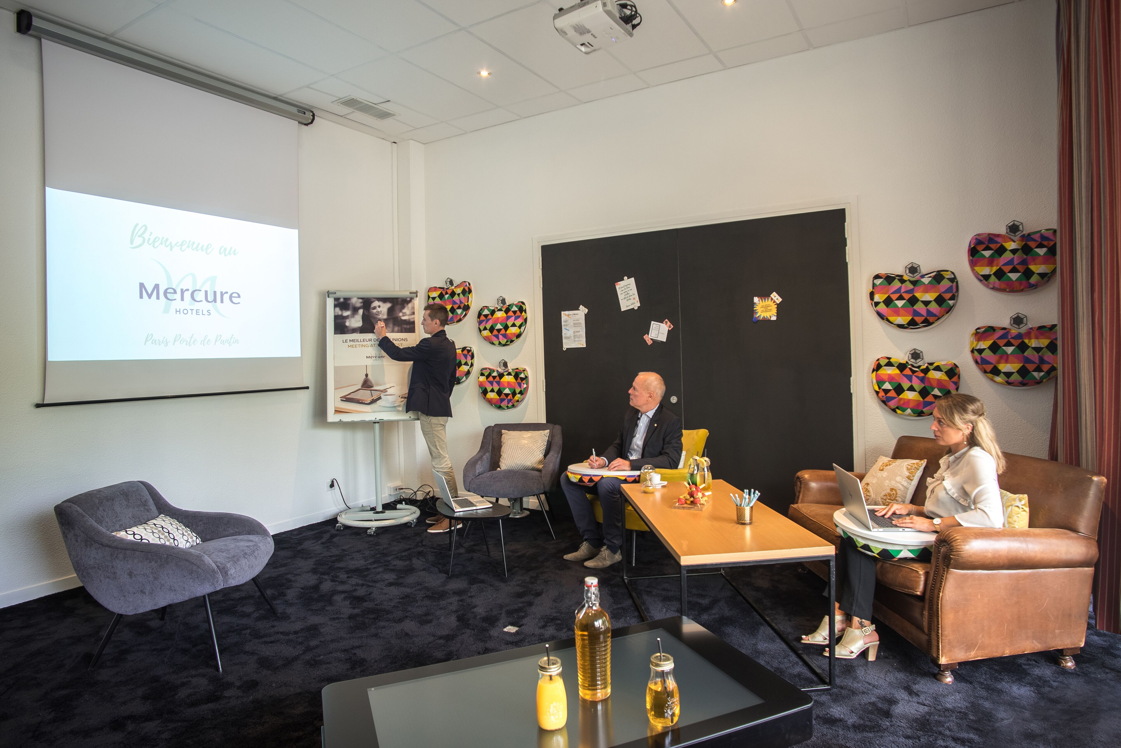 A conference room with a presentation screen, a projector, and a table with bottles. There are two chairs and a couch.