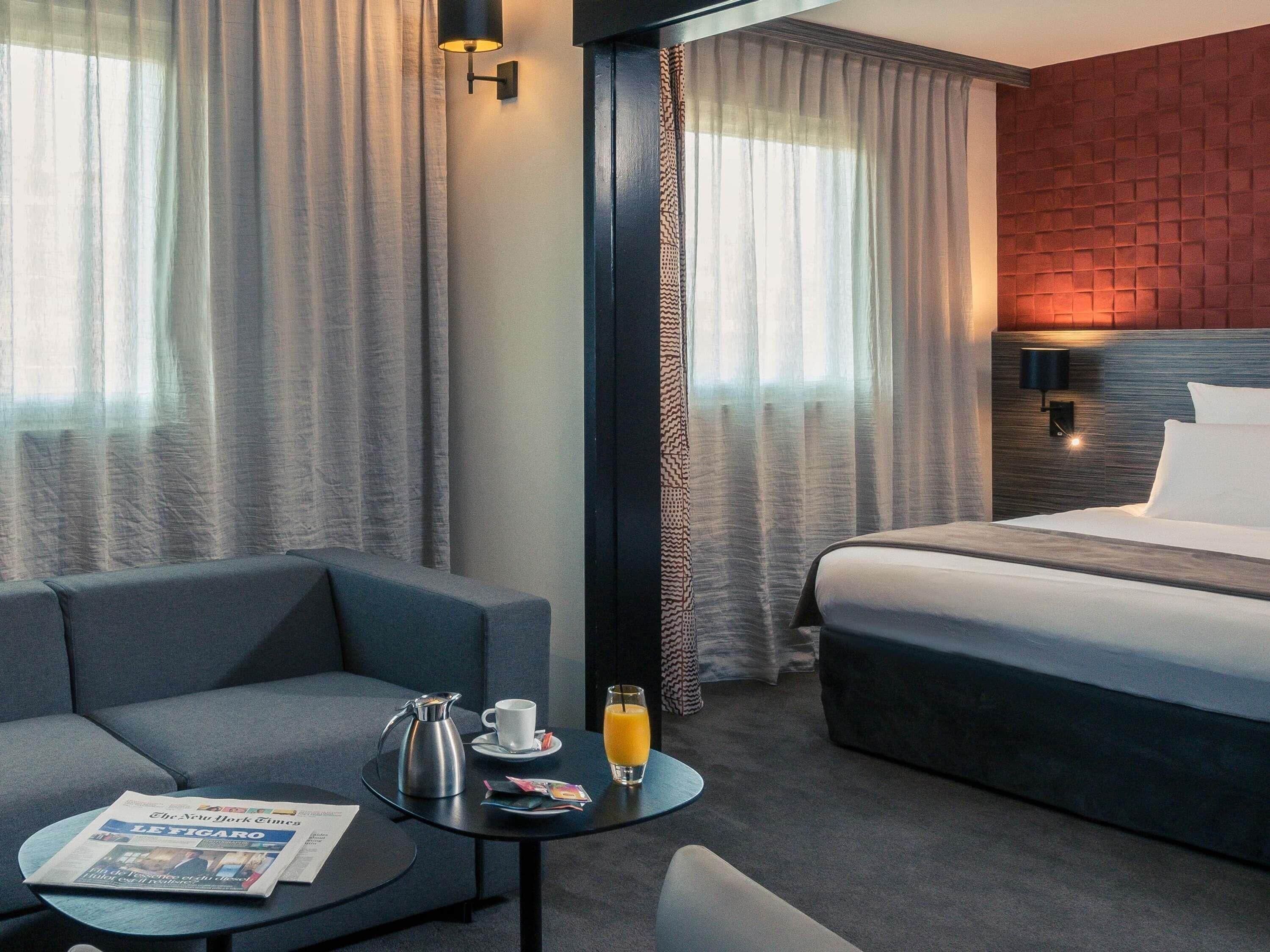 Ein Hotelzimmer mit Bett, Sofa, einem kleinen Tisch mit einer Zeitung, einer Kaffeemaschine und einem Glas Orangensaft.