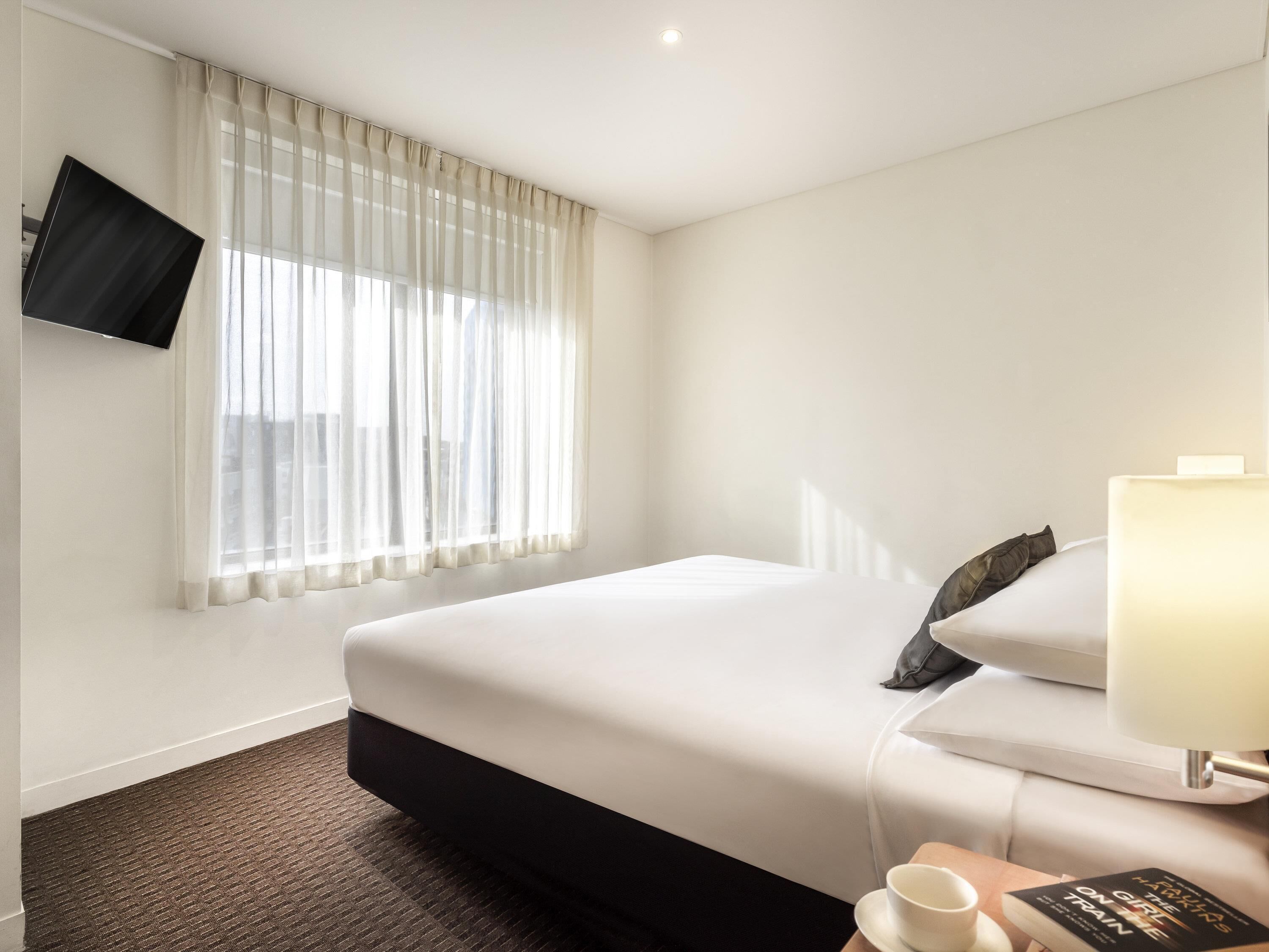 Habitación de hotel con una cama, un televisor de pantalla plana en la pared, una mesita de noche con lámpara y una ventana con cortinas translúcidas.
