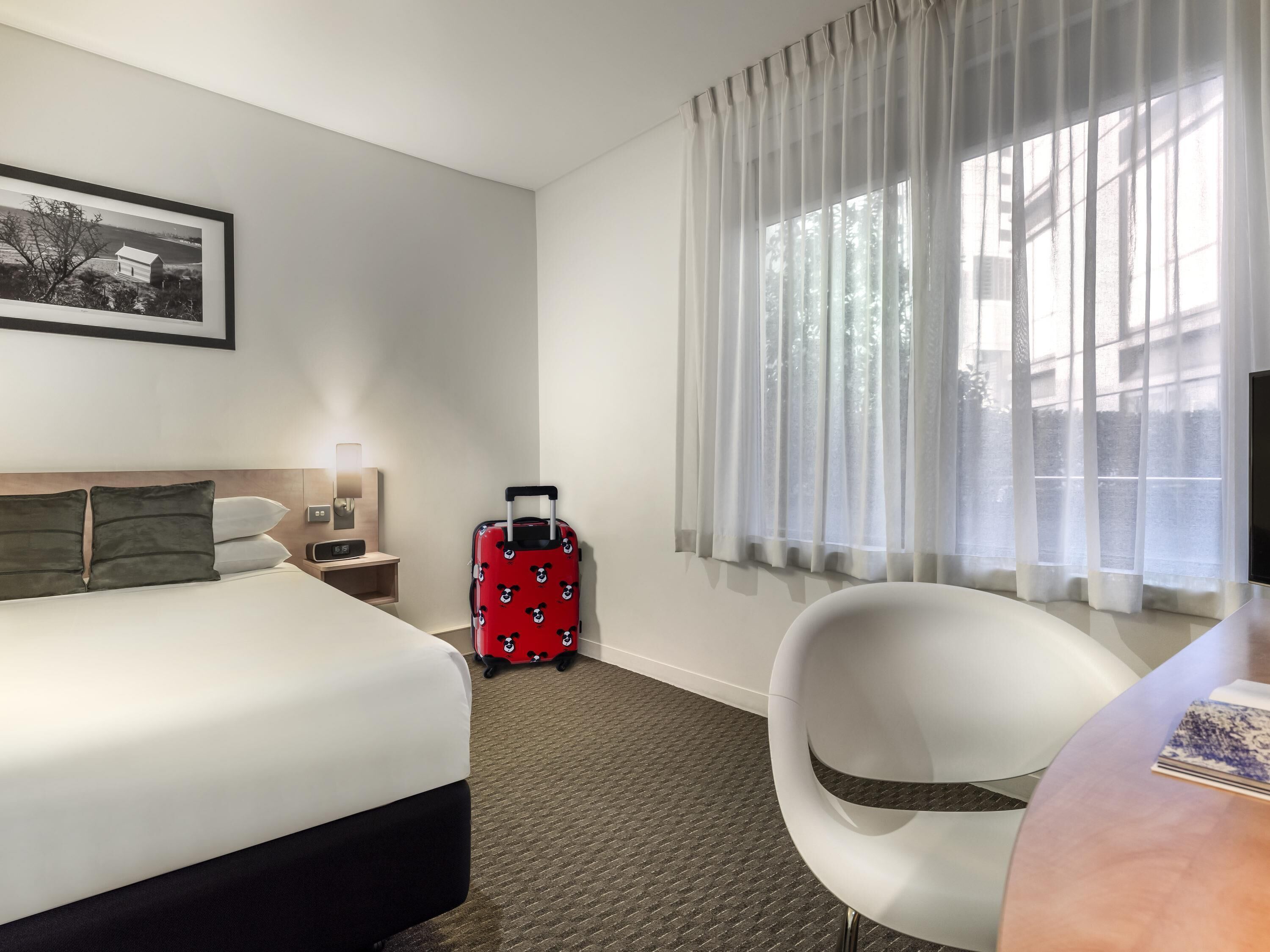 A hotel room with a bed, a chair, a suitcase, a nightstand, a lamp, and a window with curtains.