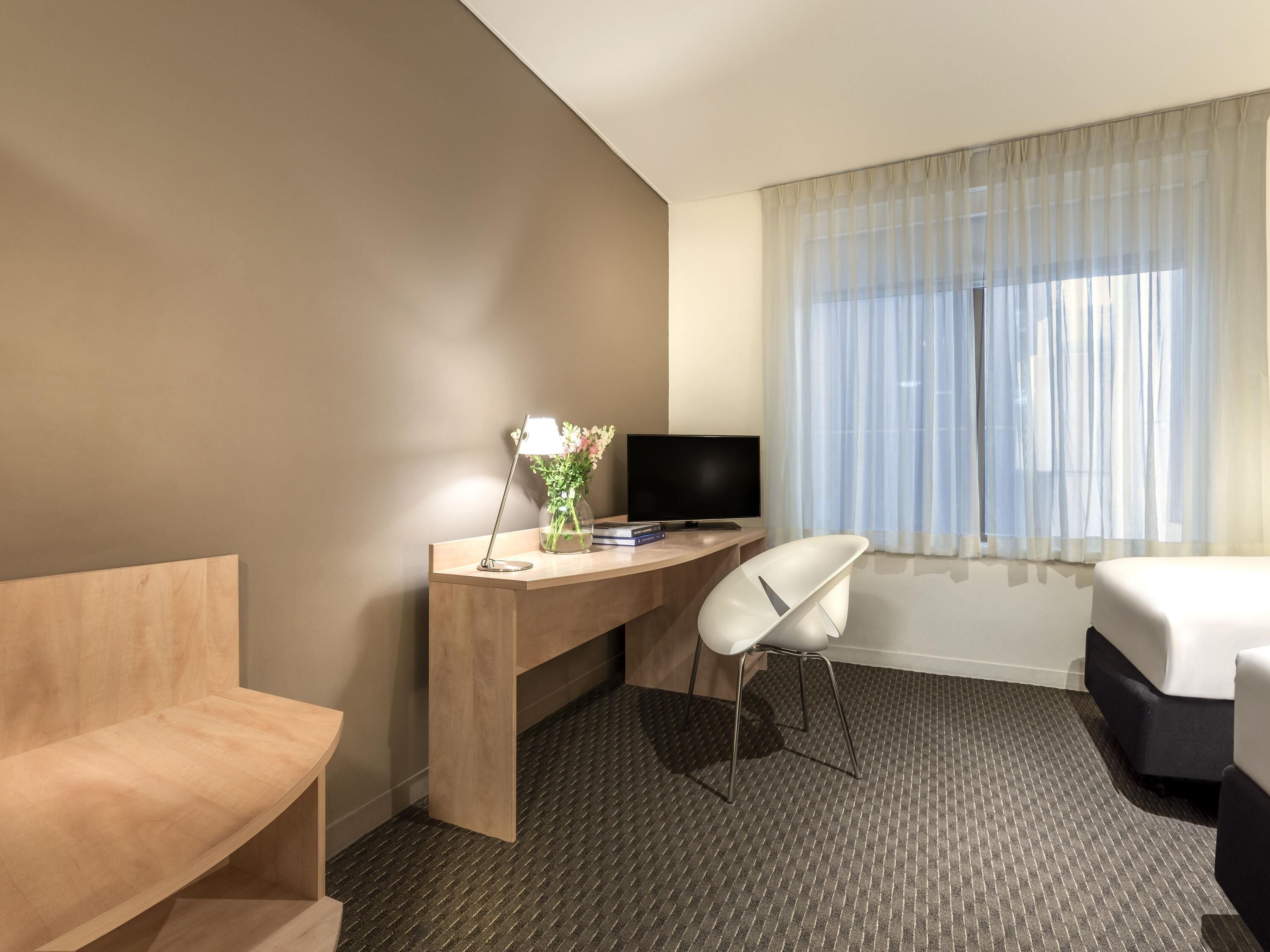 Habitación de hotel con cama, escritorio con ordenador, silla y ventana con cortinas translúcidas.