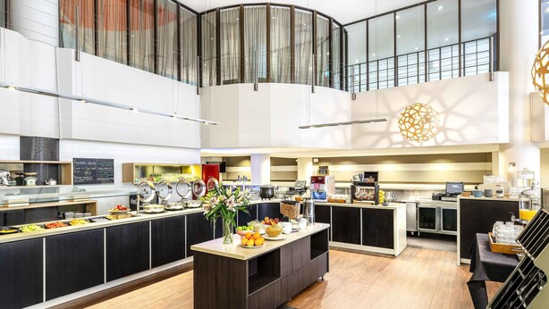 Un vestíbul d'hotel modern amb zona de buffet, racó de cafè i espai d'estar amb una escala.