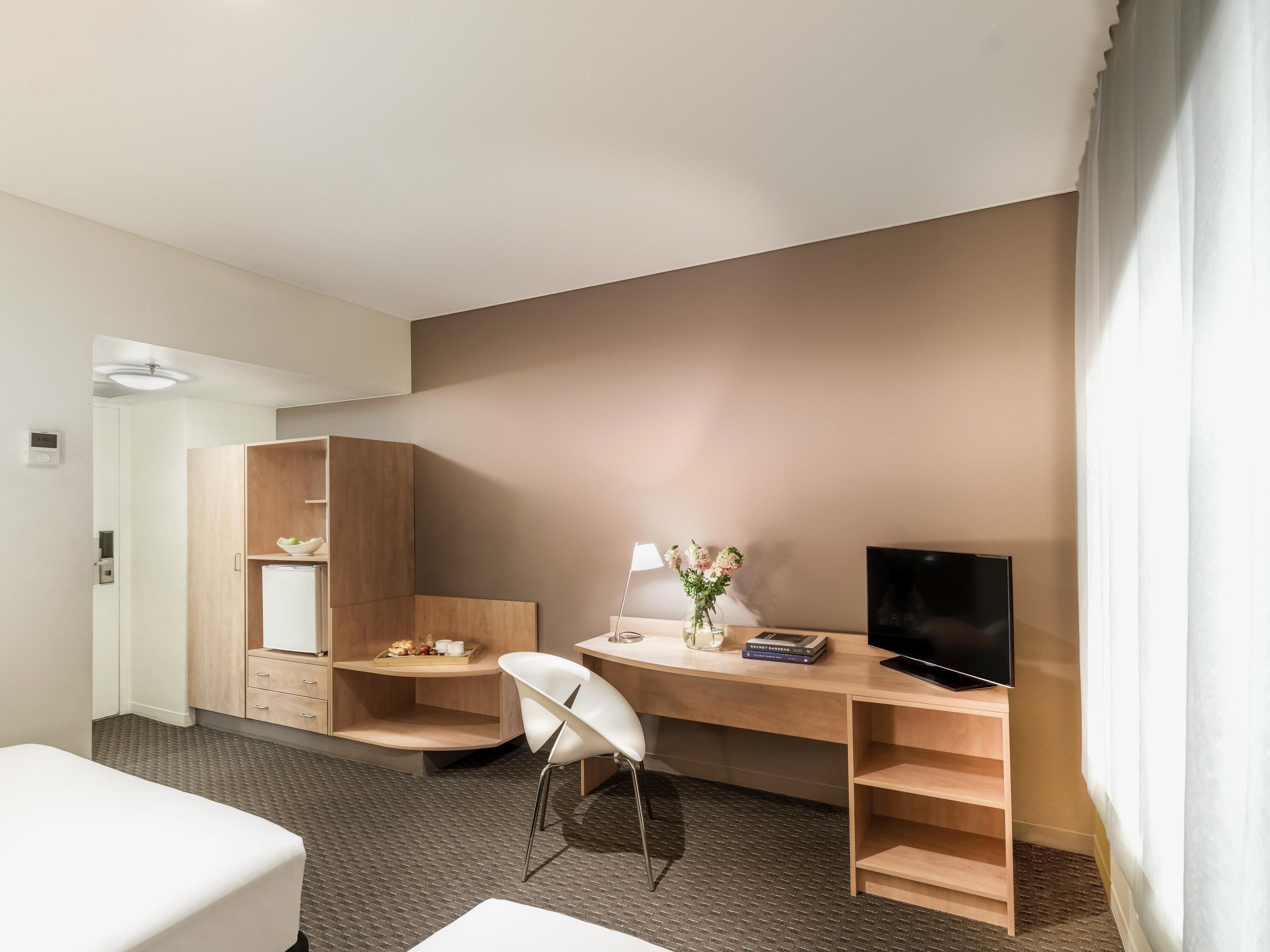 Habitación de hotel con cama, escritorio con televisor, silla, lámpara y estante con libros y un jarrón de flores.