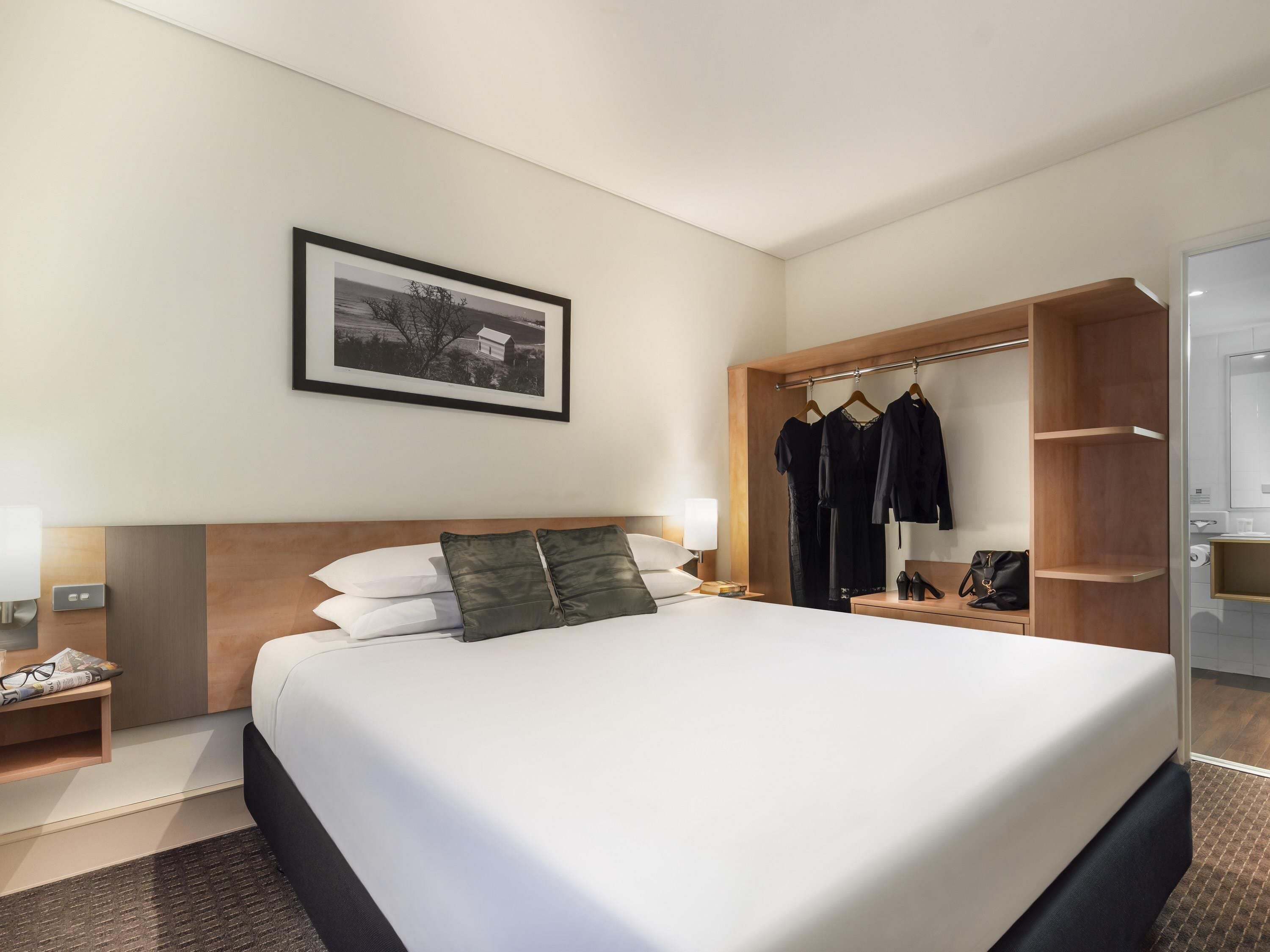 A hotel room with a large bed, a bedside table with a lamp, a wardrobe with hanging clothes, and a framed picture on the wall.