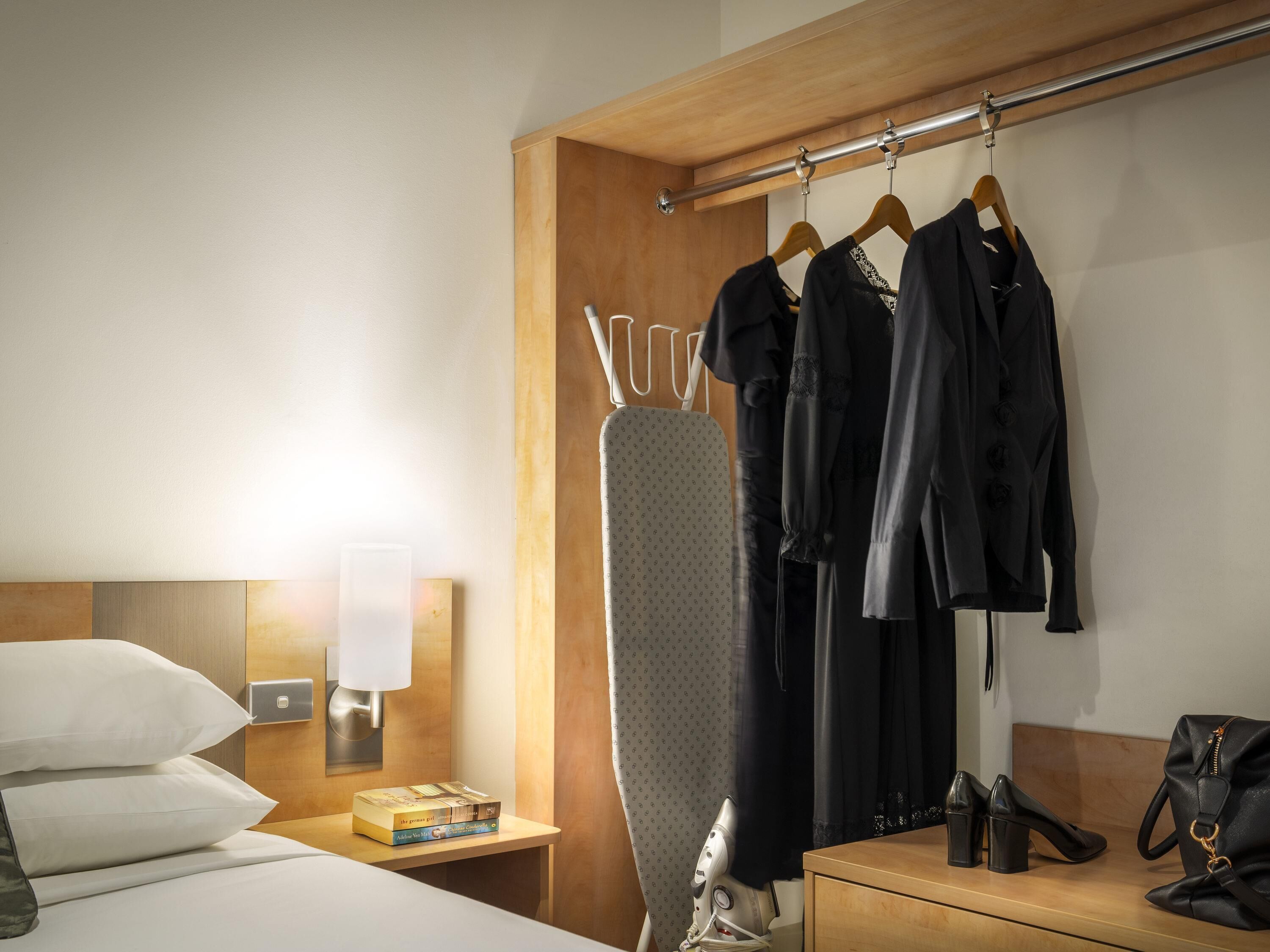 A hotel room with a neatly made bed, a nightstand with a lamp, an ironing board, a wardrobe with hanging clothes, a small table with books, and a handbag.