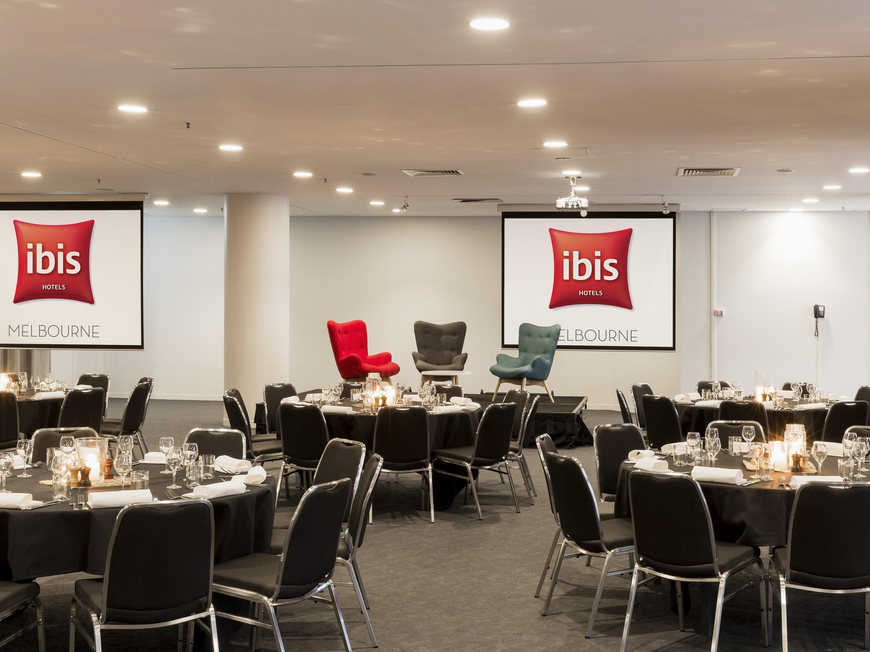 A conference room with tables set for dining, black chairs, and two screens displaying the ibis hotels logo.