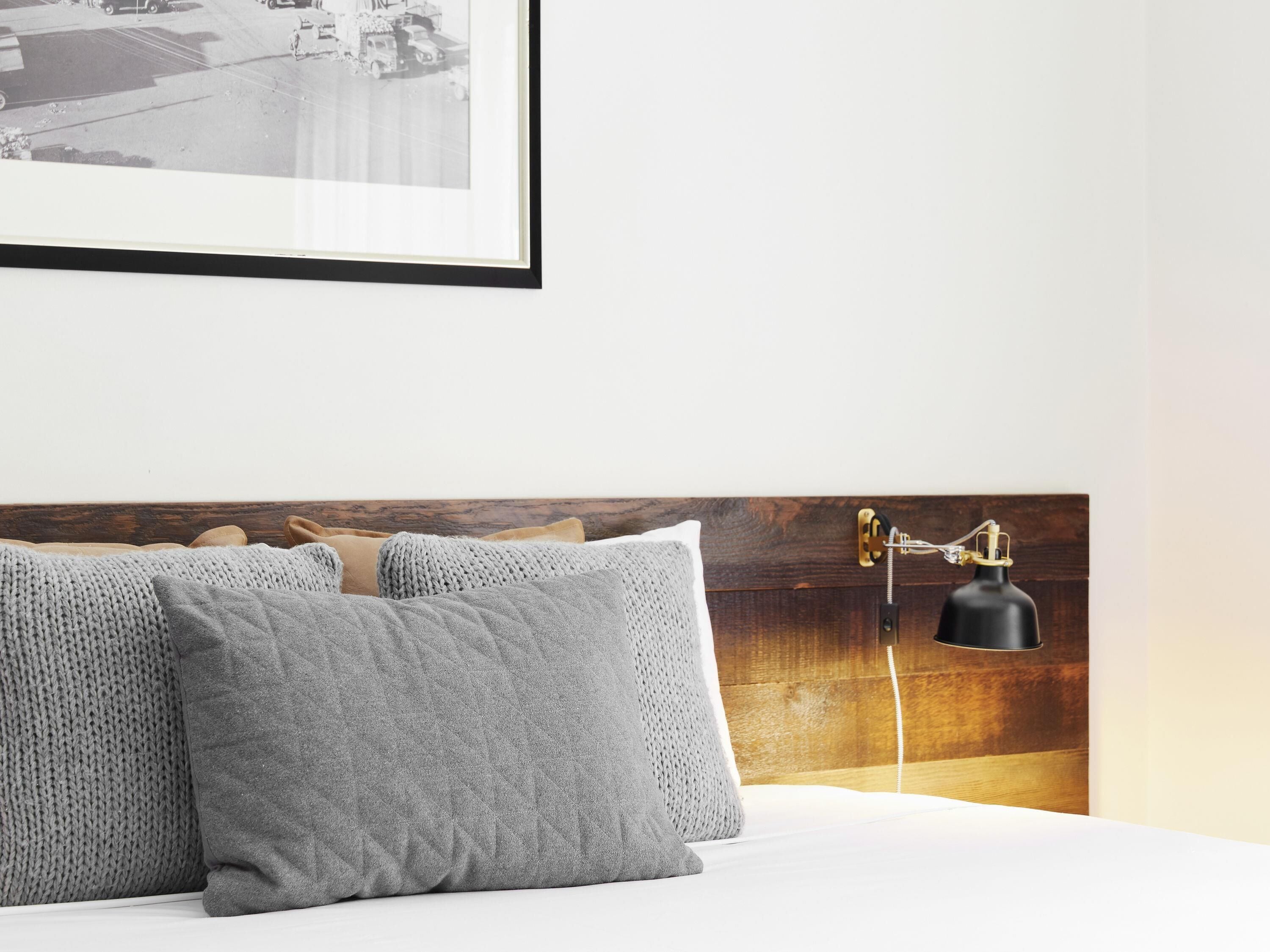 Una cama bien hecha con almohadillas grises, un cuadro enmarcado encima y una lámpara de noche.