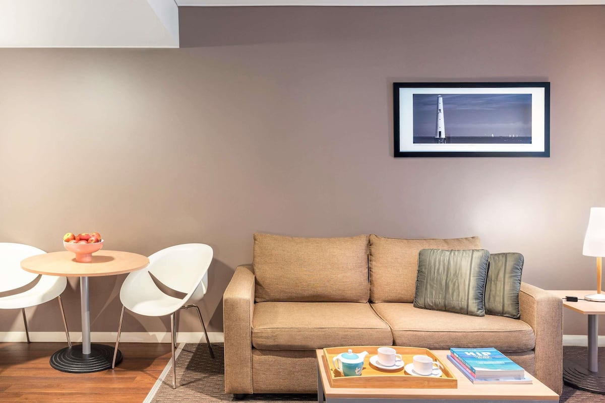 Una habitación de hotel moderna con un sofá color beige, una pequeña mesa redonda con sillas, una mesita con una bandeja de té y un cuadro en la pared.