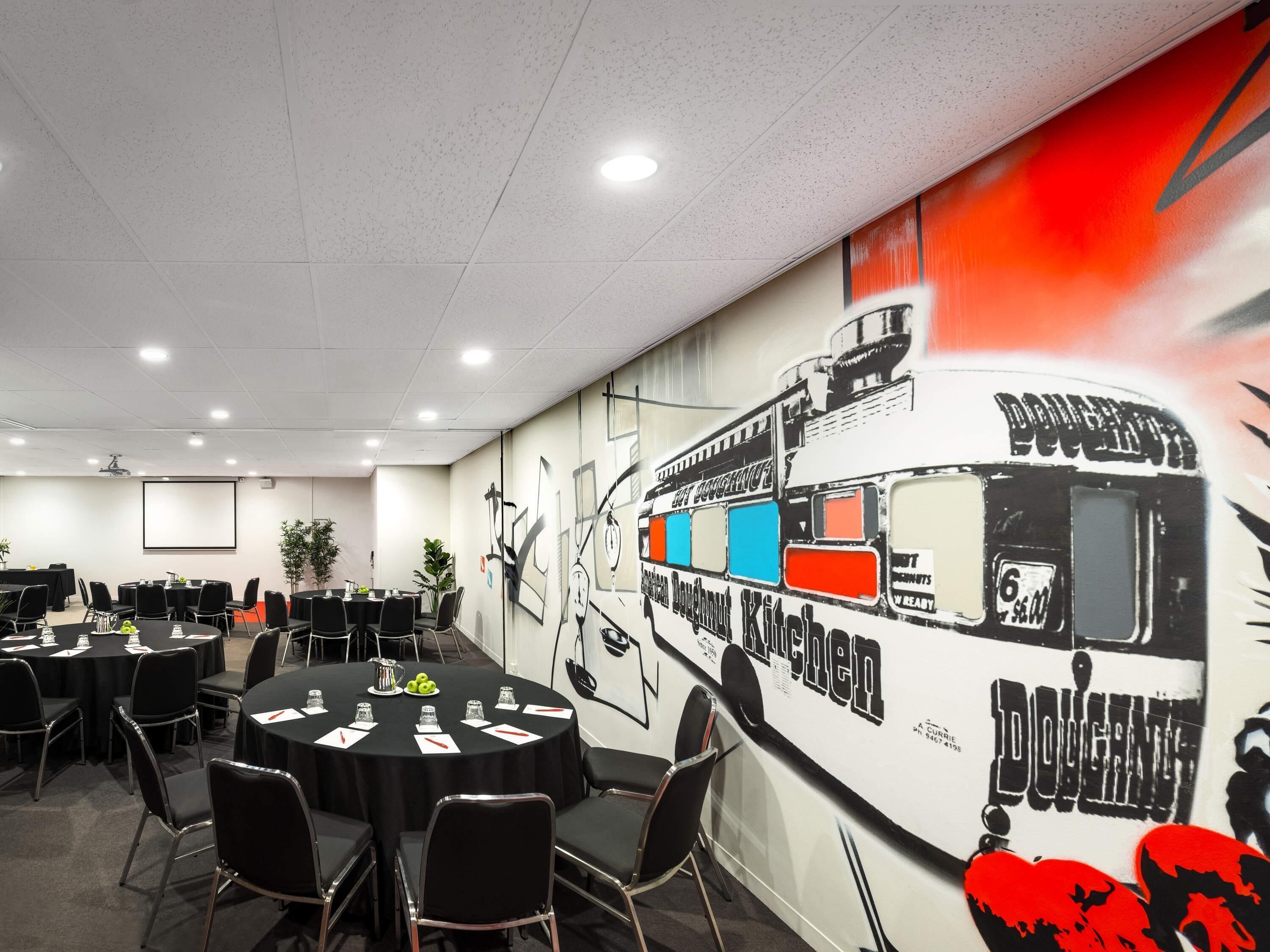A conference room with tables, chairs, a projector screen, and a mural of a doughnut truck.