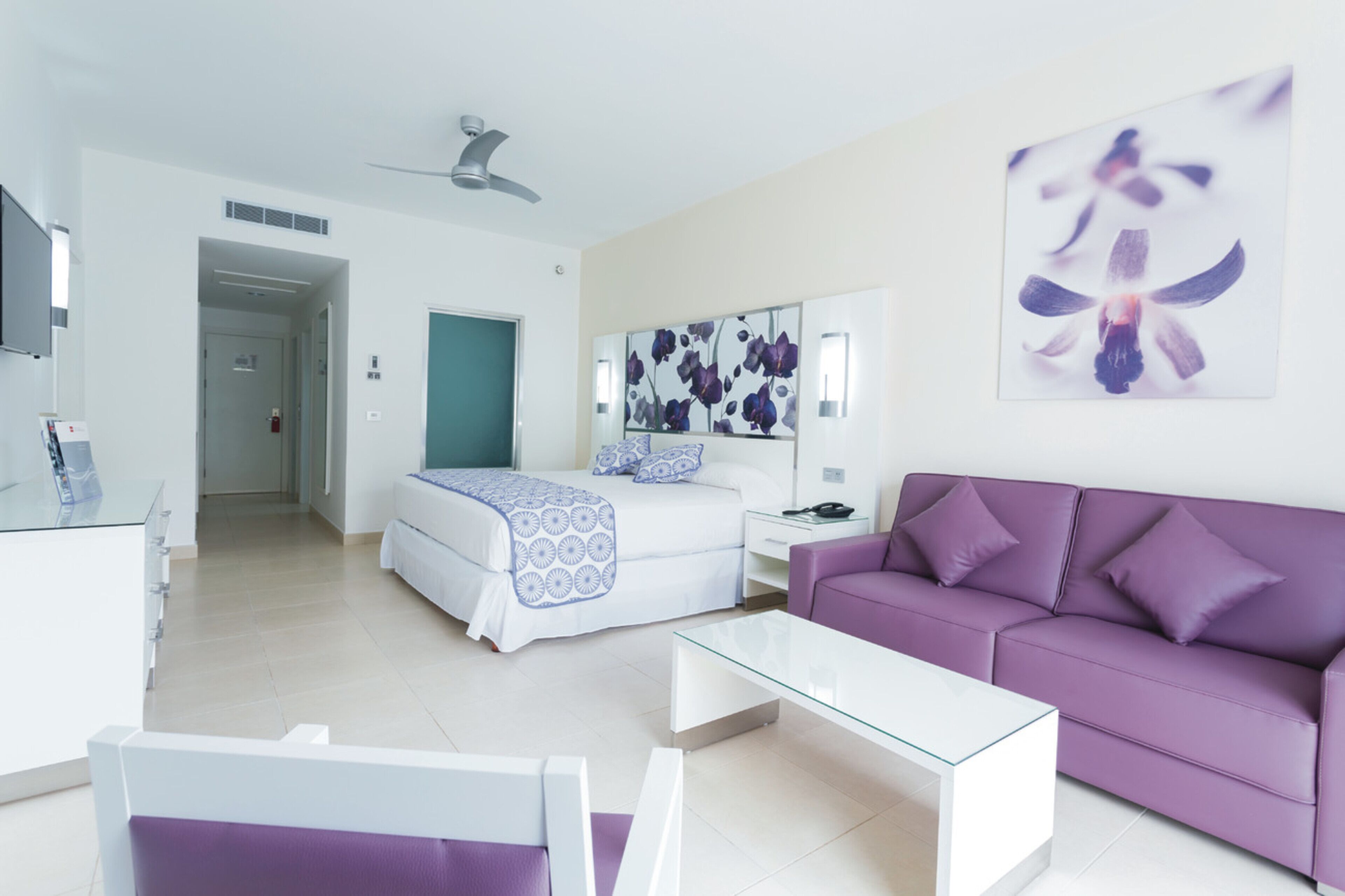 Una camera d'hotel moderna con un letto, un divano viola, un tavolino in vetro e un'opera d'arte appesa al muro.
