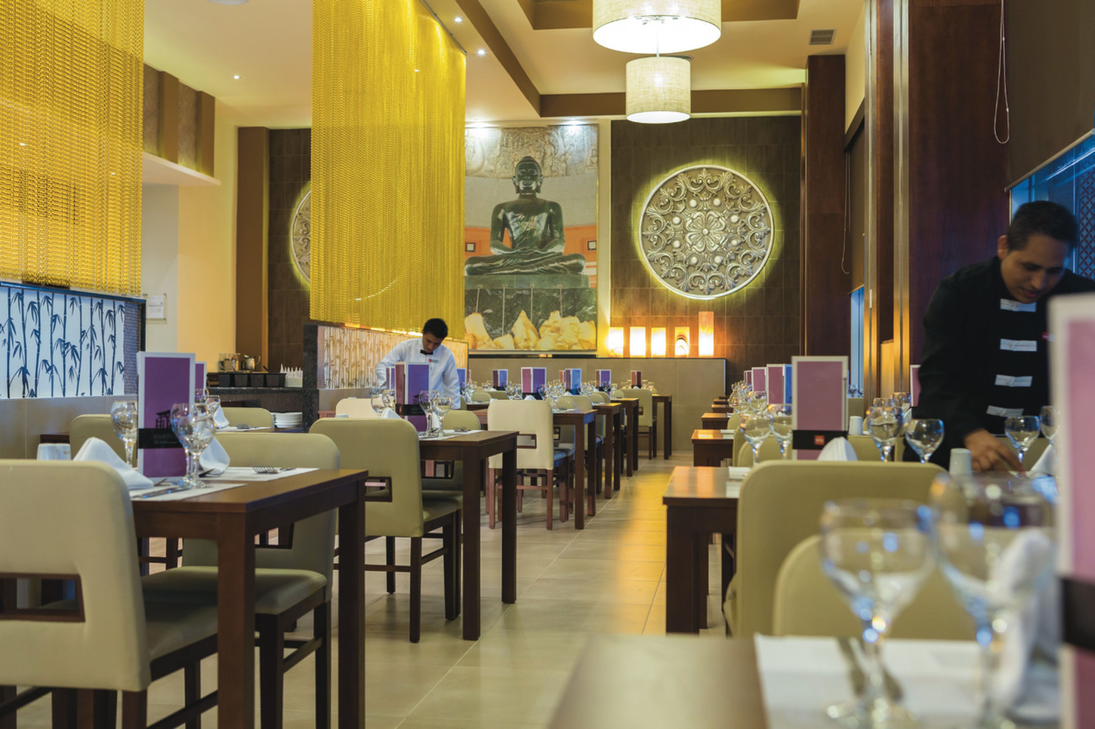 A restaurant with multiple tables, a large Buddha statue, and a waiter attending to guests.