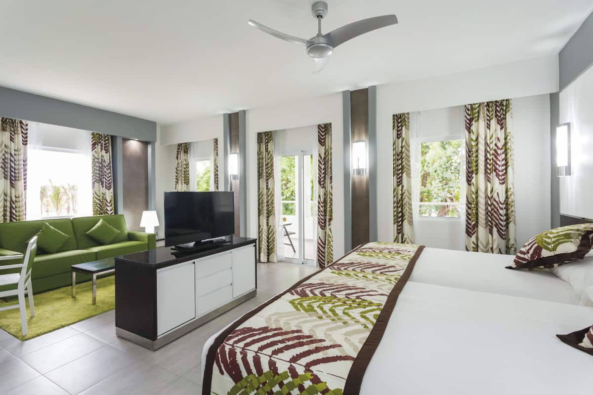 Quarto de hotel moderno com uma cama grande, um sofá verde, uma televisão e uma varanda com cortinas.