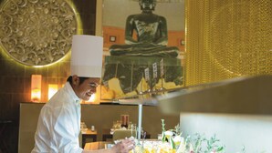Un chef, vêtu d’un uniforme blanc, prépare un dessert, avec une statue de Bouddha en arrière-plan.