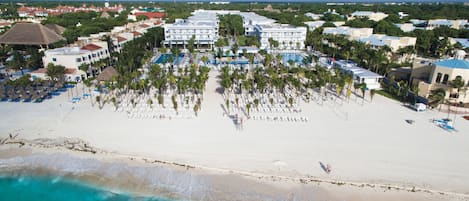 An aerial view of a beach resort with a large pool area, numerous palm trees, and a sandy beach.