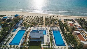 Vue aérienne d’un complexe hôtelier doté de deux grandes piscines, d’un accès à la plage et de palmiers.