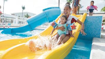 Un tobogán de agua en una zona de piscina, con un tobogán azul y amarillo, palmeras y una valla blanca al fondo.