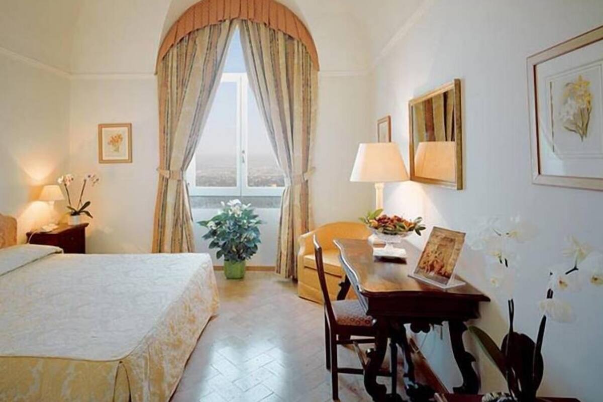 Een hotelkamer met een bed, een bureau met stoel, een kamerplant en een raam met gordijnen.