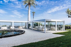 Un área de comedor exterior moderna con paredes de vidrio, una palmera y un pequeño estanque.