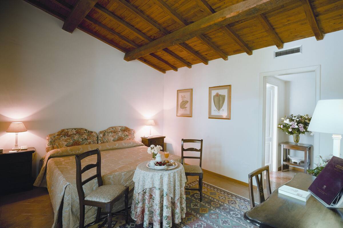 Een gezellige slaapkamer met een houten plafond, een bed met bloemenbeddengoed, twee stoelen, een klein rond tafeltje met een bloevas en een nachtkastje met een lamp.