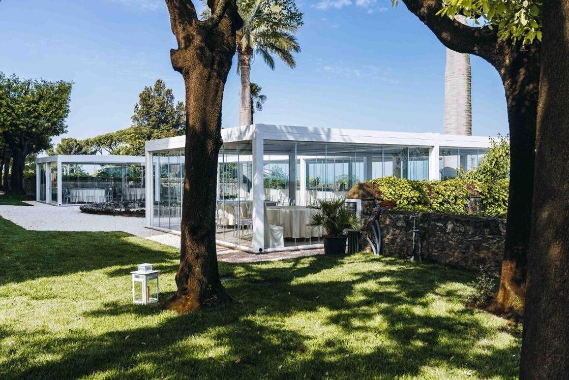 A modern glass structure with outdoor seating, surrounded by trees and a stone wall.