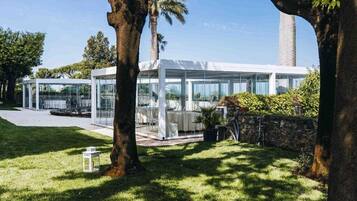 A modern glass structure with outdoor seating, surrounded by trees and a stone wall.