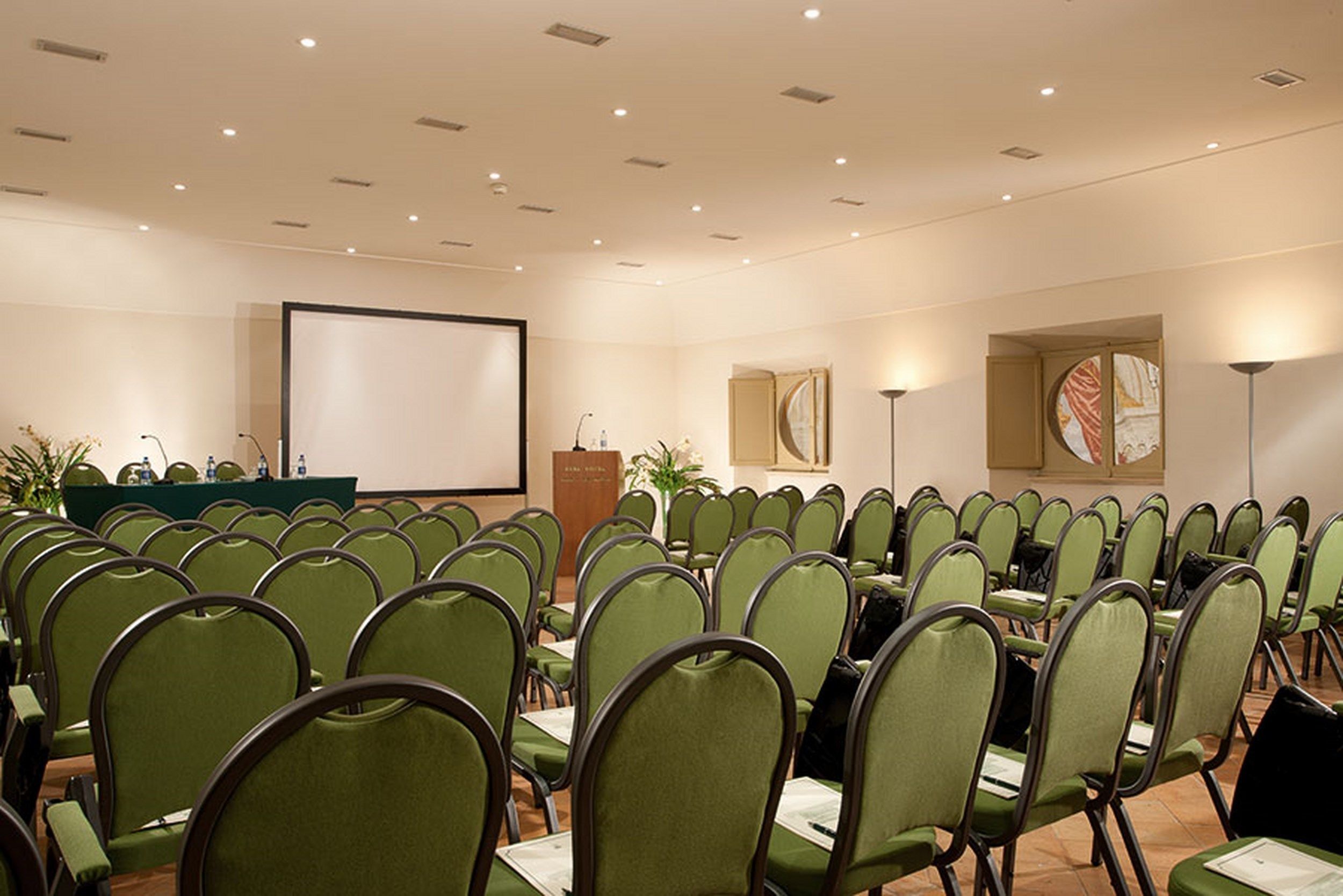 A conference room with rows of green chairs, a podium, a large screen, and wall art.
