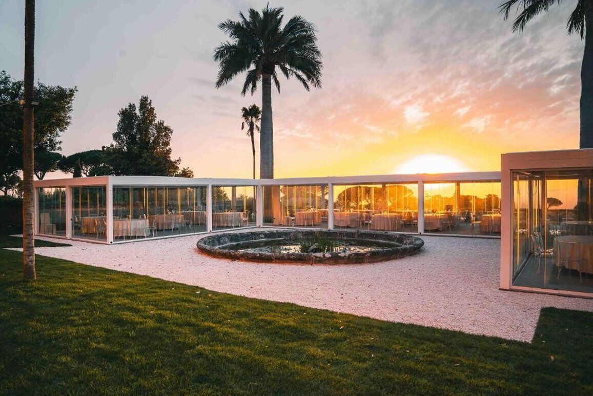 A modern outdoor dining area with glass enclosures, a circular garden, and palm trees.