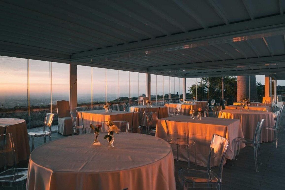 Ein Außenbereich zum Essen mit runden Tischen und Stühlen, großen Fenstern und Blick auf die Landschaft.