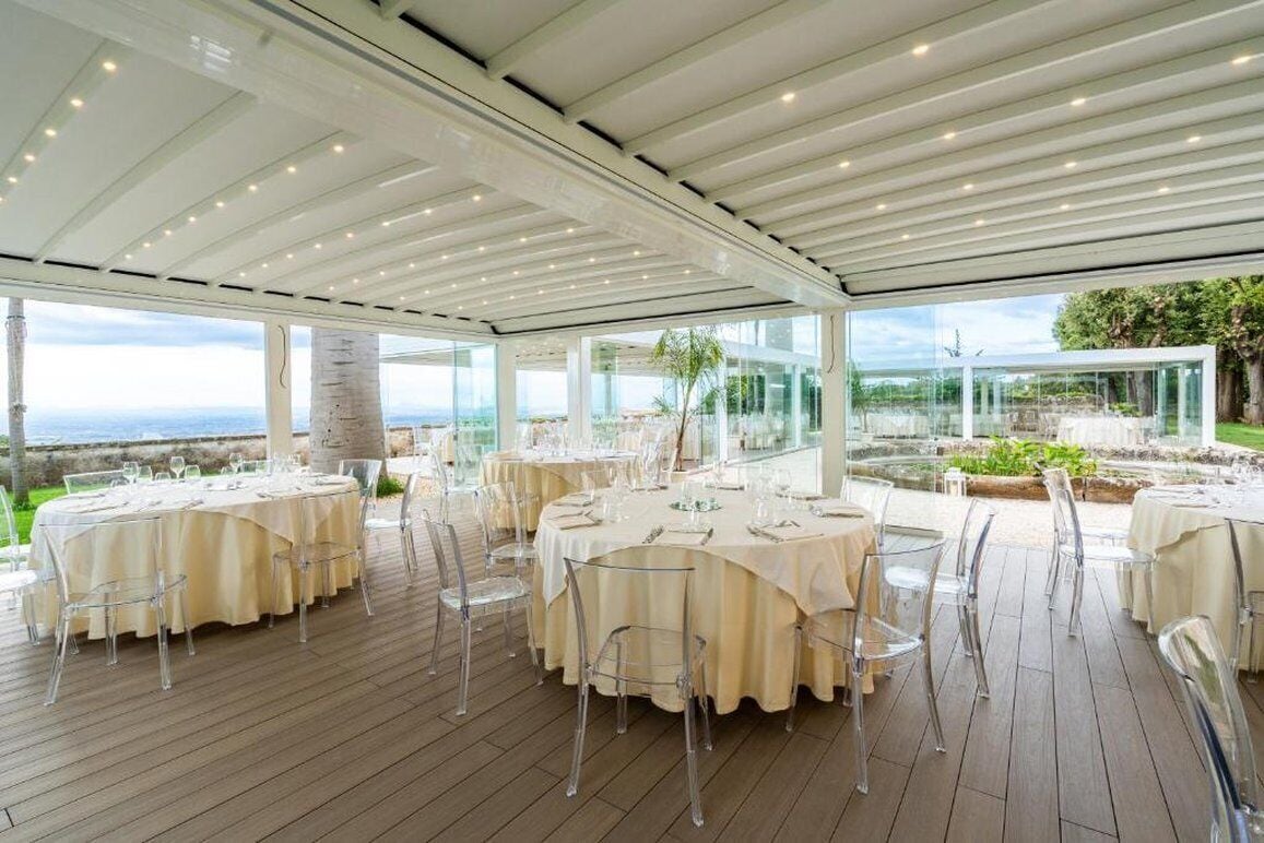An outdoor dining area with round tables, clear chairs, and a view of the sea.