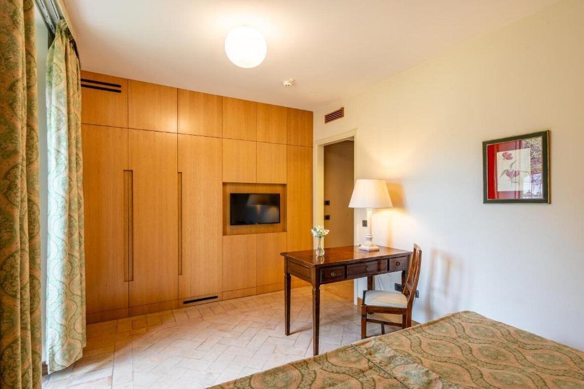 Habitación de hotel con armario de madera, escritorio y silla, cama con cubrecama estampado, televisor montado en la pared y un cuadro enmarcado.