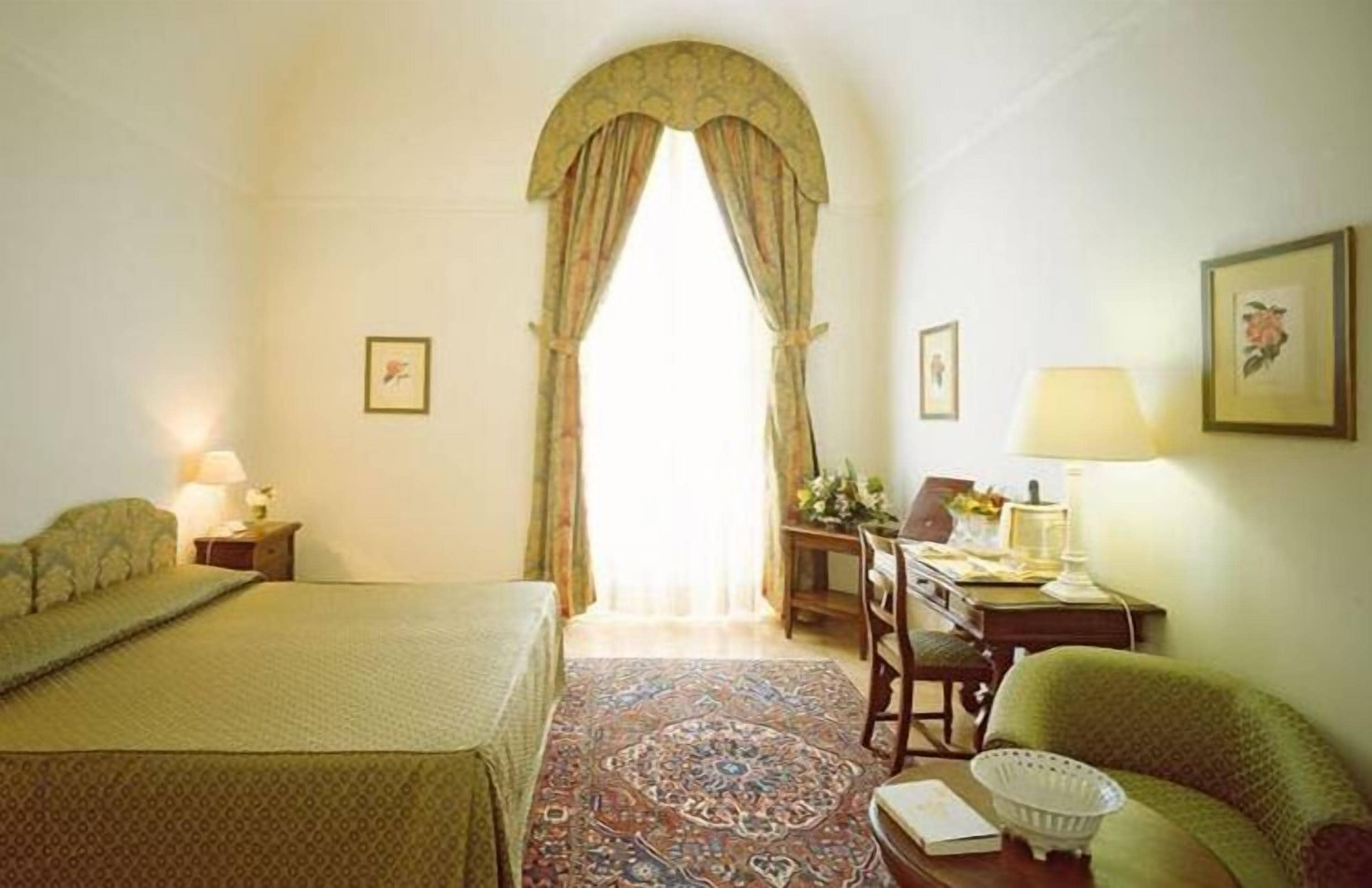 Una habitación con una cama, un escritorio, una silla, una lámpara y una ventana con cortinas.
