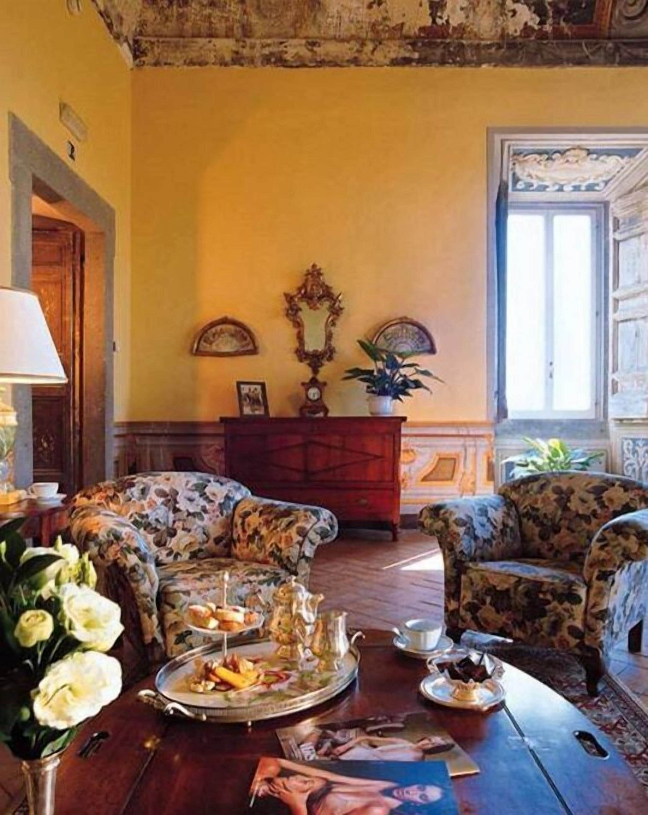 A room with floral-patterned armchairs, a wooden cabinet, and a dining table with a tray of food.
