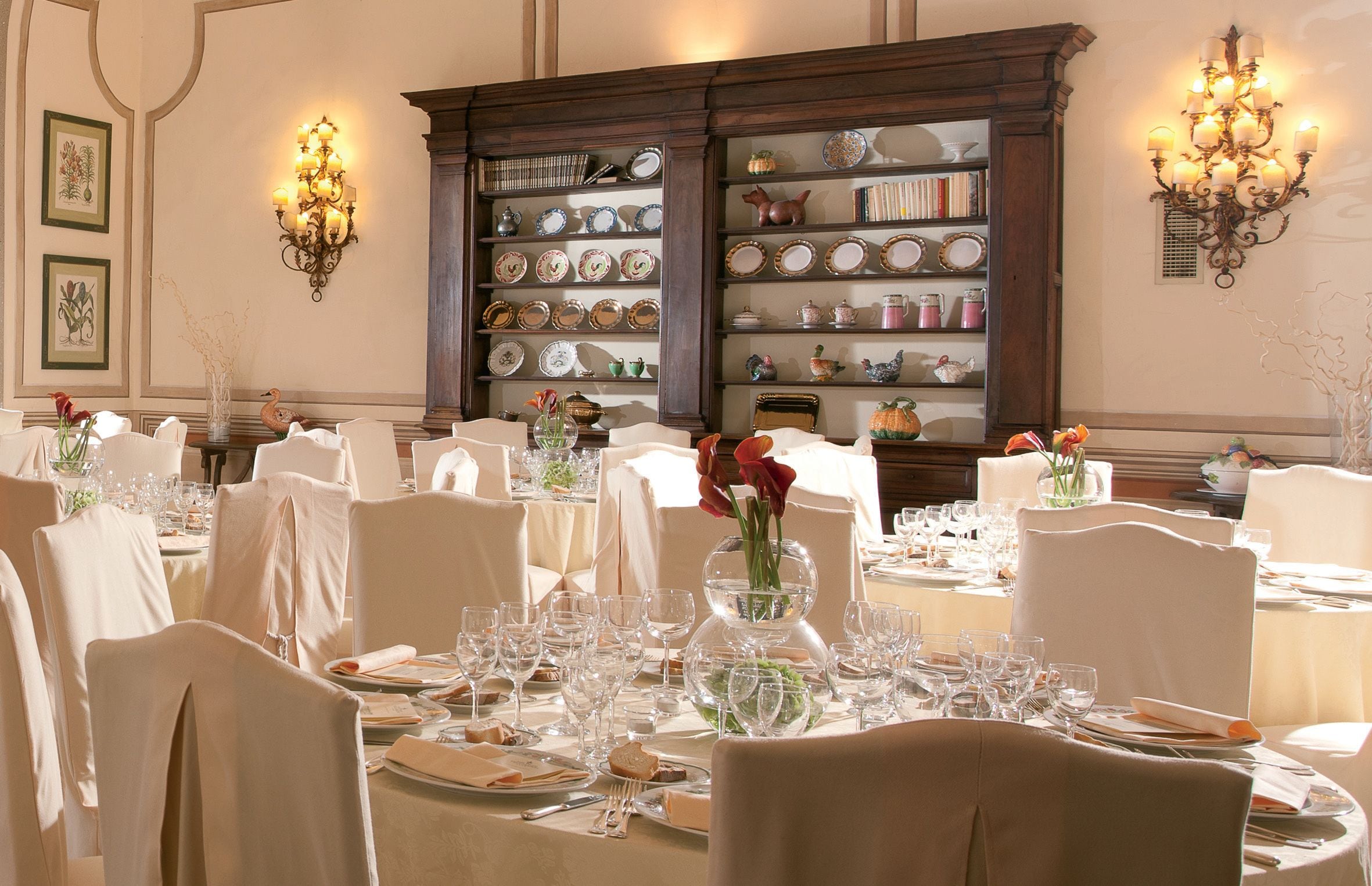 A formal dining room with round tables set for a meal, white chairs, and a wall display of china and glassware.