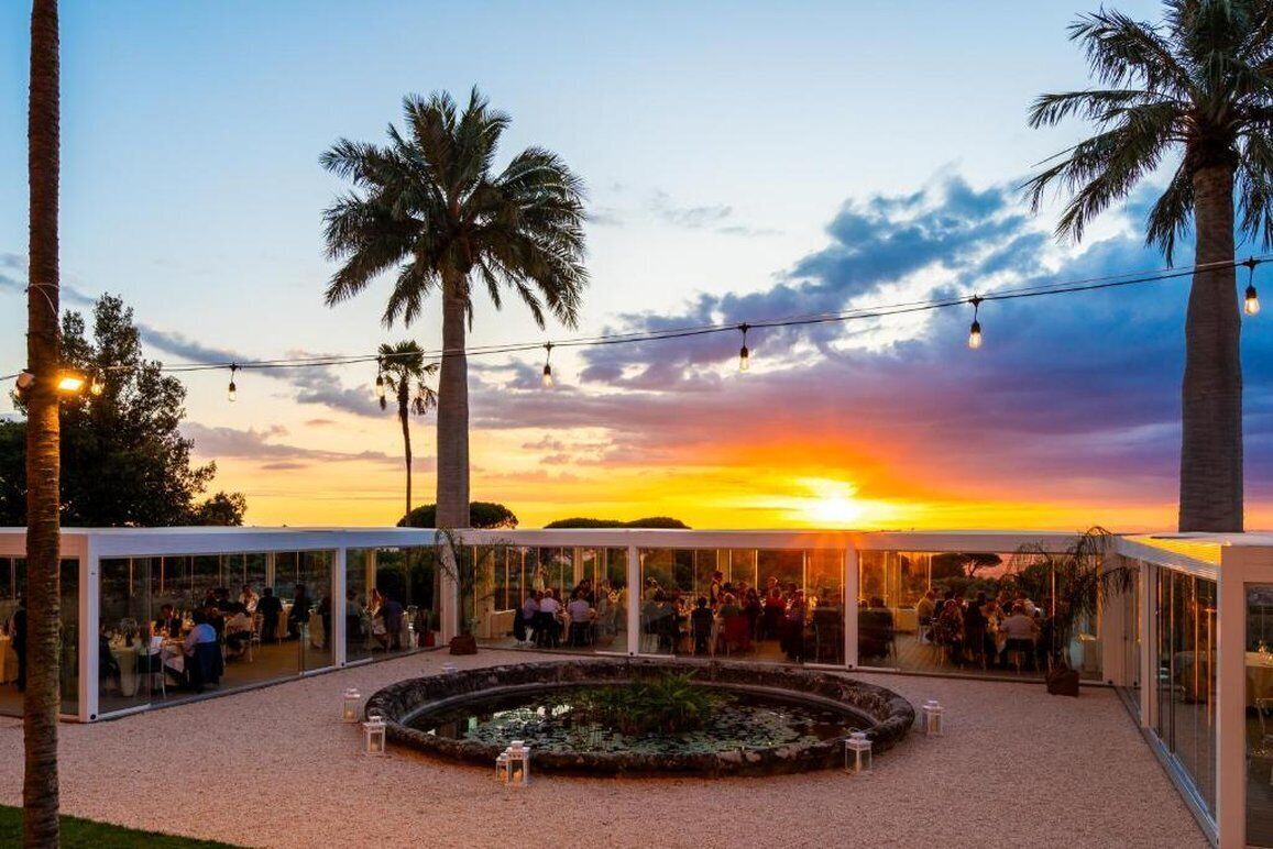 Un espace de restauration en plein air agrémenté de palmiers, d’une fontaine circulaire et de guirlandes lumineuses, le tout se détachant sur le ciel au coucher du soleil.