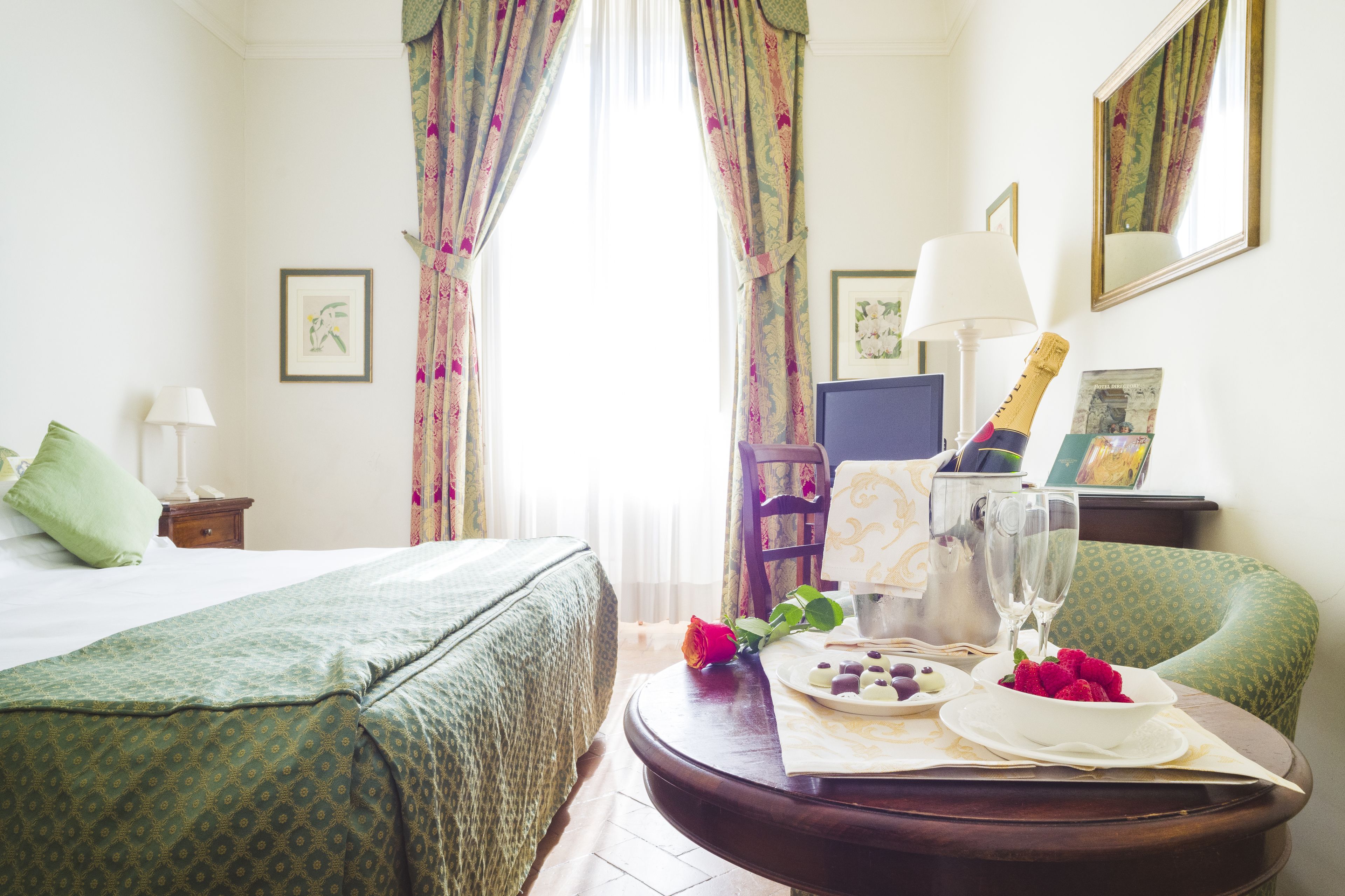 A hotel room with a bed, a dining table with a bottle of champagne, glasses, and a plate of fruit.