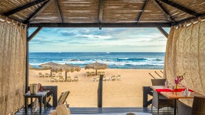 Un restaurant situé en bord de mer, offrant une vue sur l’océan, doté de parasols en chaume et d’une table dressée pour le dîner.