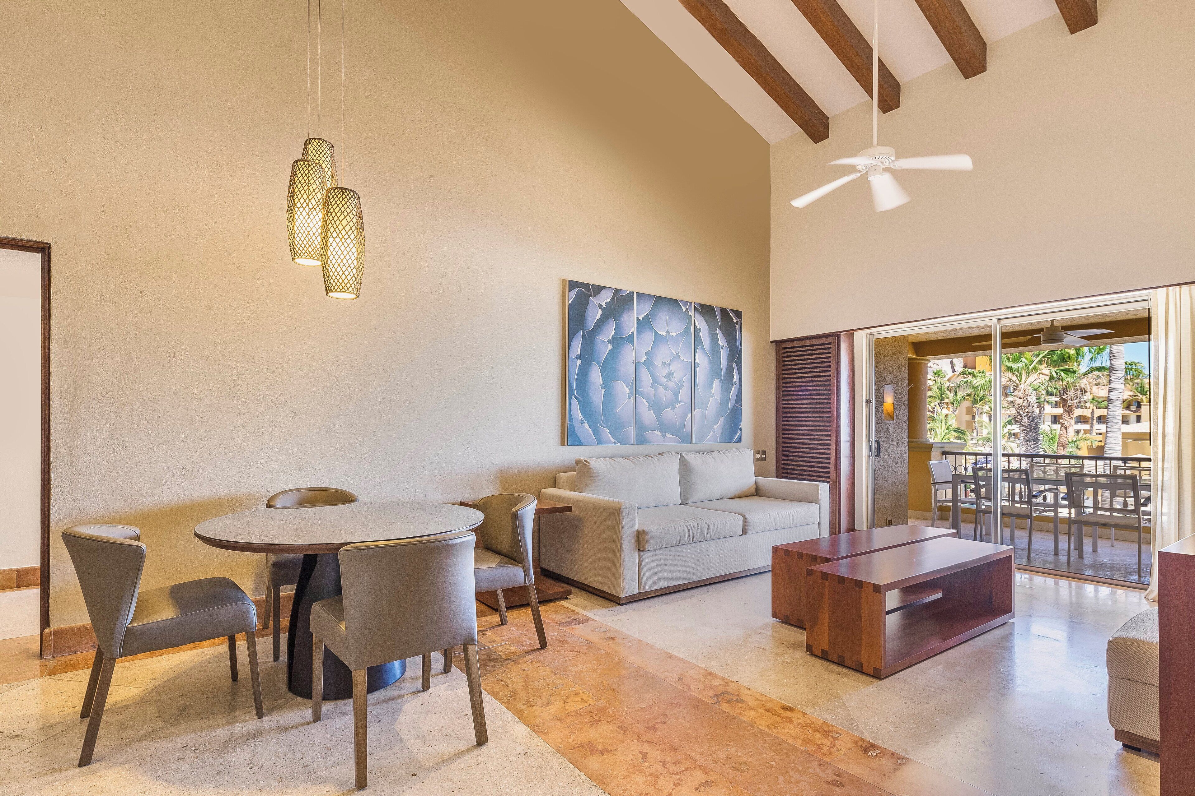 A modern living room with a round table, grey chairs, a sofa, and a wooden coffee table. There is a ceiling fan and a large abstract painting on the wall.