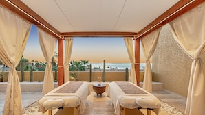 Une salle de spa équipée de deux tables de massage, d’une petite table avec une bougie et offrant une vue sur l’océan.