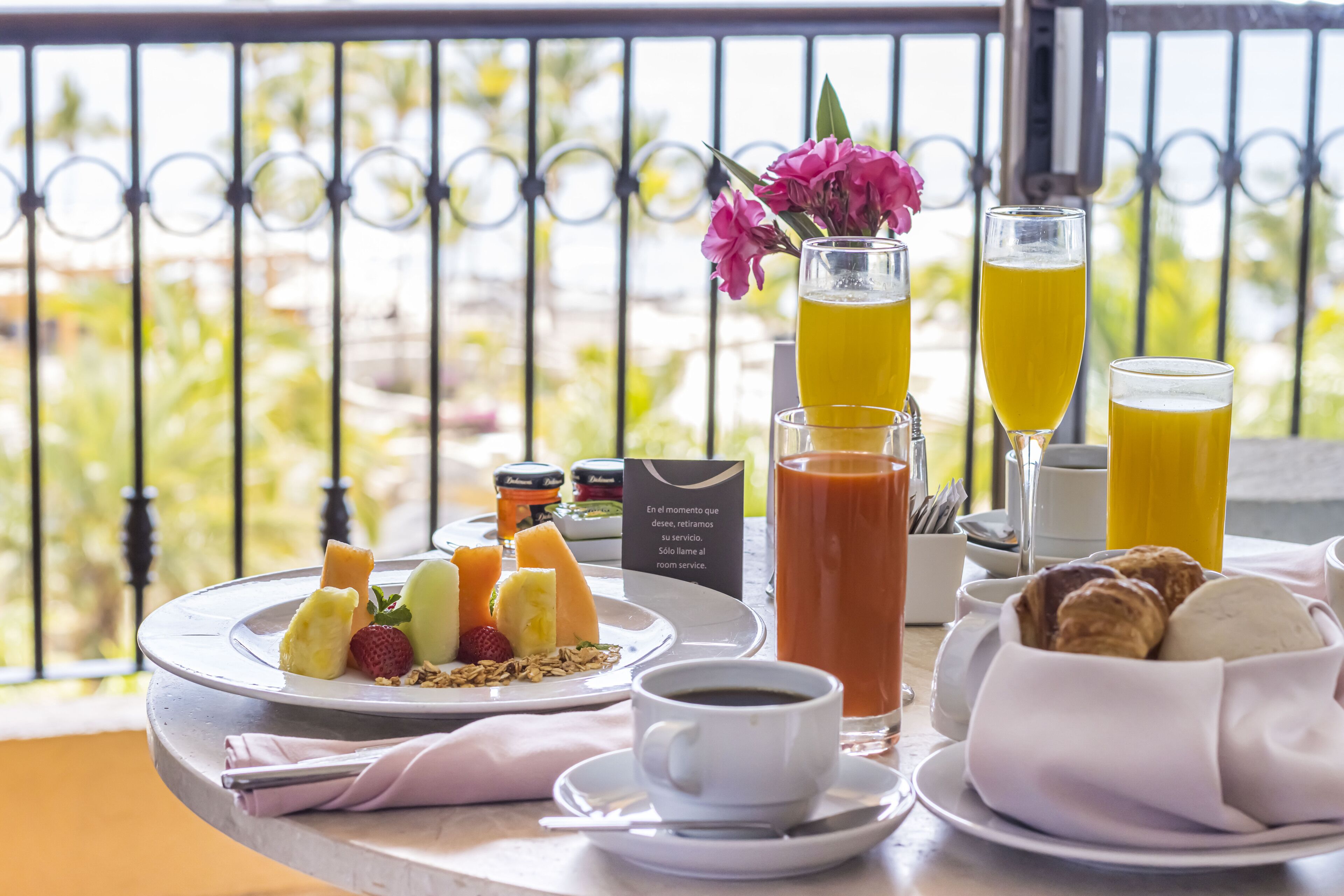 A breakfast setting with a variety of fruits, pastries, and beverages on a balcony overlooking a scenic view.